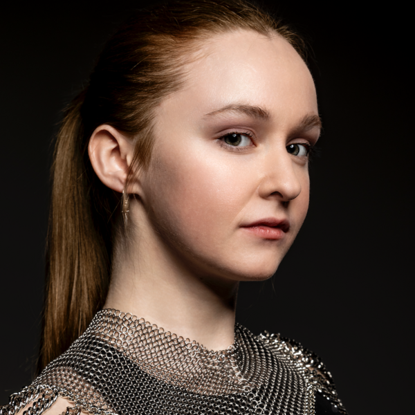 A white fem presenting person's headshot. she looks at the camera at a 3/4 angle. her dark blonde hair is pulled into a ponytail, and she is wearing a black tank top, small dangling silver knife earrings, and a silver chainmail neck piece. her expression is neutral, and her eyebrows are raised.