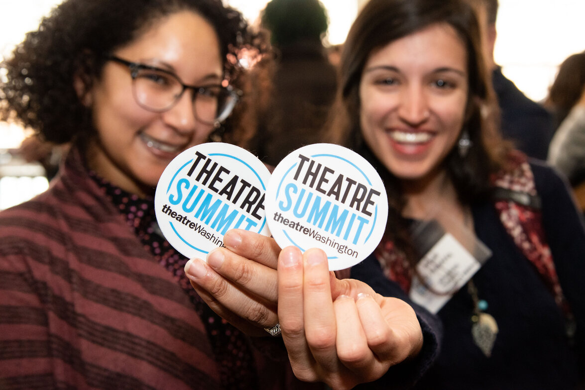 two women holding up "theatre summit" white circular stickers