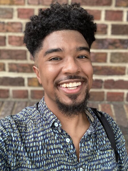 Headshot for Connor Lugo-Harris, a medium-skinned  black man with curly hair in a green geometric patterned shirt smiling in front of a brick wall