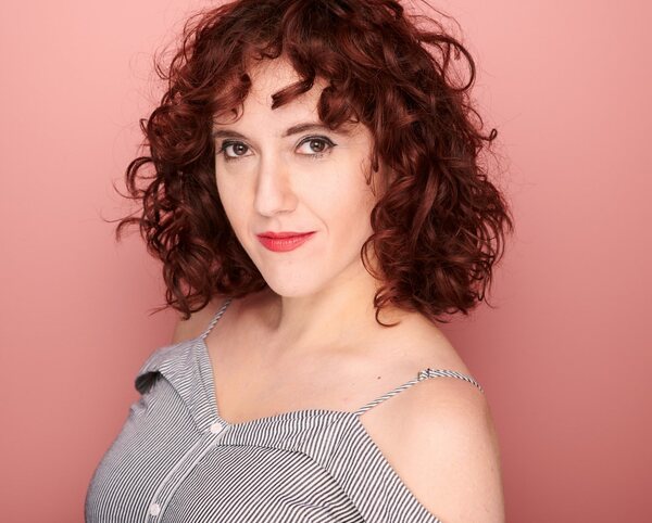 a white woman with red curly hair in a strapless striped shirt, against a pink background