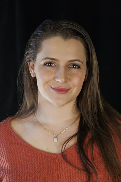 smiling white woman, young, long brown hair, brown eyes, orange quilted shirt, gold necklace with pink pendant.