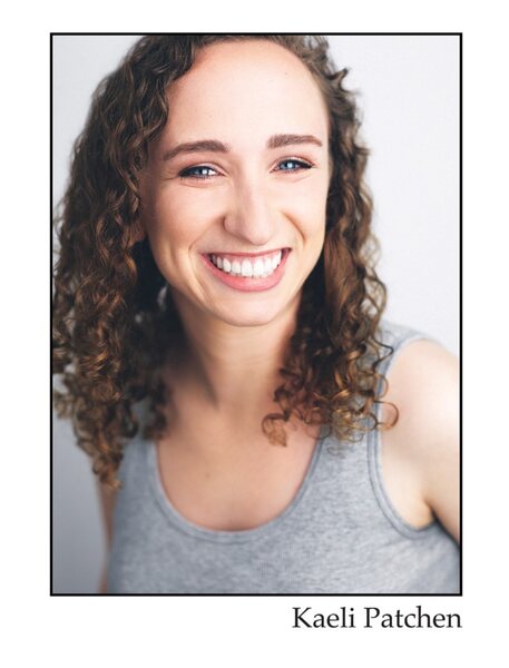 A white, brunette woman with curly hair in a gray tank top smiling in front of a white background