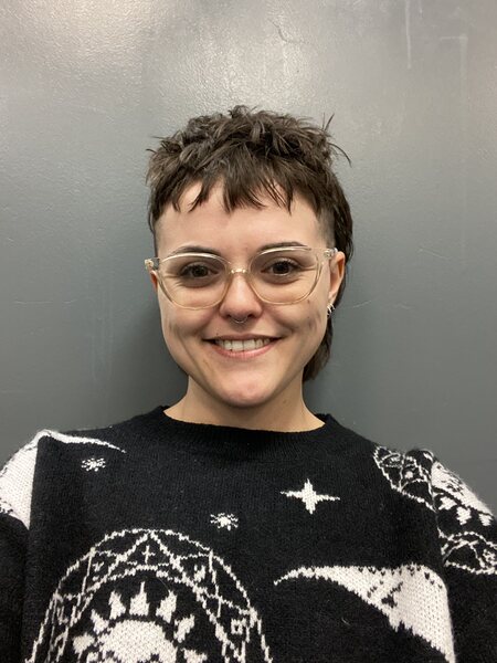 white woman with clear glasses and brown mullet hair. They are against a gray wall and is wearing a black sweater with white details on it. 