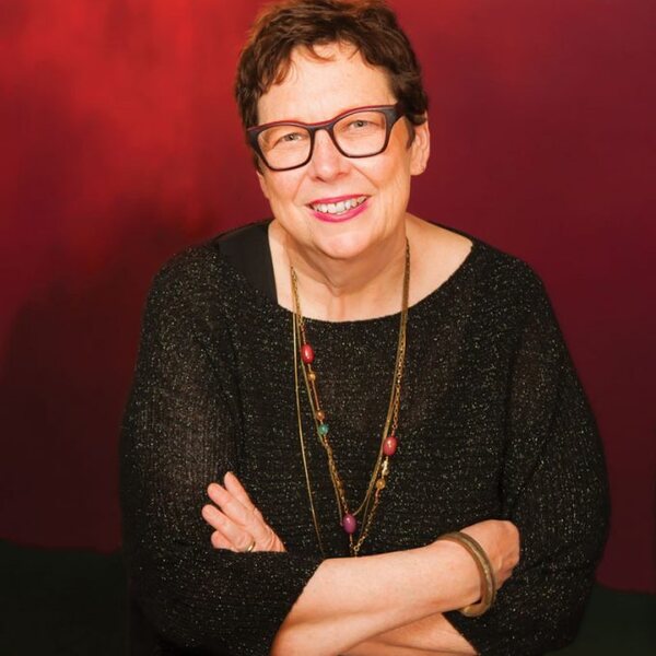 White woman wearing glasses, black dress, and colored necklaces with a short haircut in front of a red backdrop