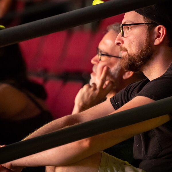 Justin A. Miller, scene designer, sits next to the director during technical rehearsals. The director is an older man who wears glasses and has salt and pepper hair and a beard. Justin wears a black T-shirt and black baseball cap. They sit in the red, plush seats of an indoor audience. The photo show them discussing the set from behind audience railings. 